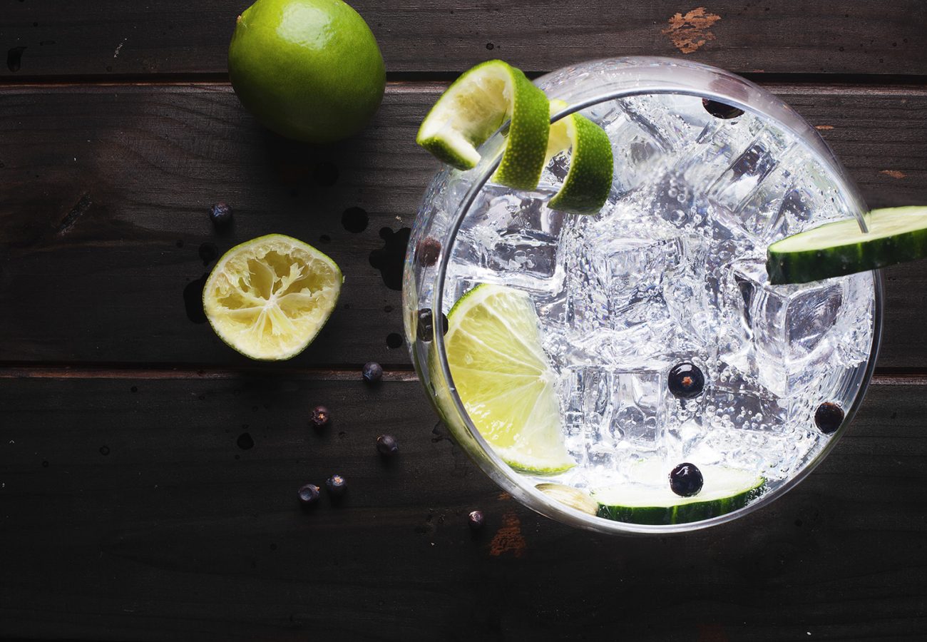 Glass of gin tonic with ice, cucumber, lime over a dark wood tab